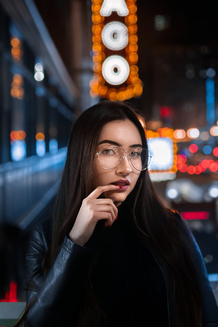Young woman in glasses posing at night with city lights backdrop and bokeh effect.