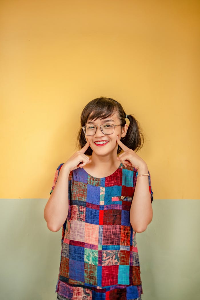 Asian woman wearing patchwork shirt and eyeglasses smiles and gestures with delight against a yellow wall.