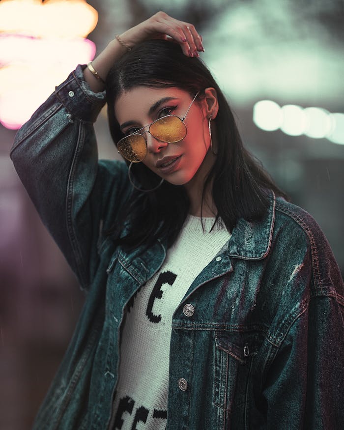 Fashionable woman posing confidently with sunglasses and denim jacket during a night shoot.
